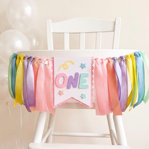 Puede incluir: Una trona blanca decorada con una pancarta que muestra la palabra ONE en letras pastel. La pancarta está adornada con un espiral amarillo, estrellas pequeñas y un fleco de cintas de satén multicolores en rosa, amarillo, morado y azul claro.