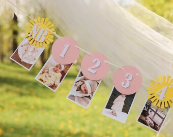 May include: A pink and yellow baby milestone banner with photos of a baby at different ages. The banner includes the numbers 1, 2, 3, and 4, and the letters "NB" for newborn.