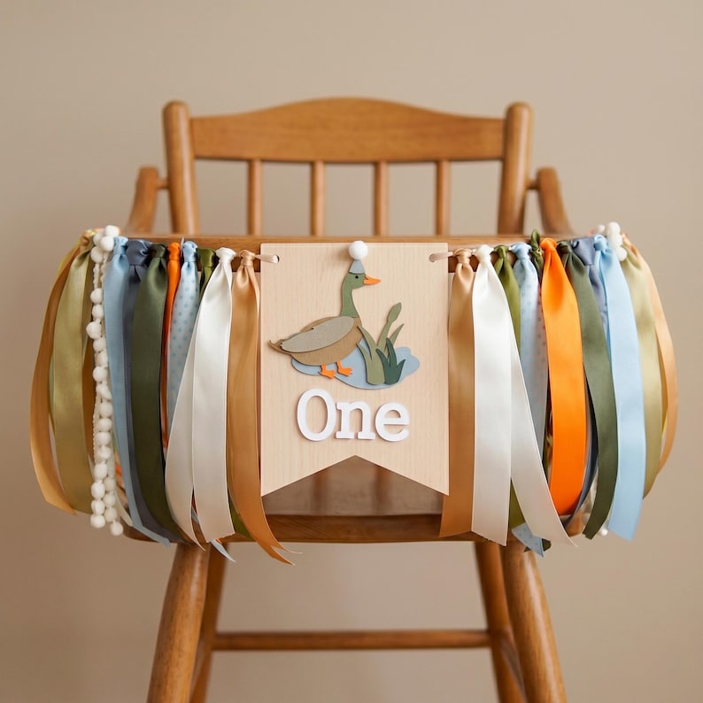 May include: A wooden high chair banner with a duck illustration and the word "One". The banner is decorated with colorful ribbons in shades of blue, green, orange, and gold. White pom-poms are also attached to the banner.