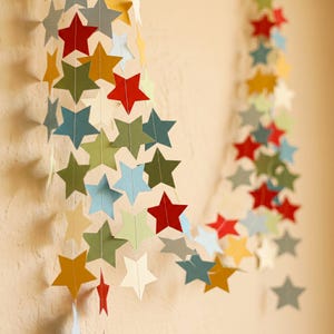 Puede incluir: Una guirnalda de estrellas de papel de colores rojo, amarillo, verde, azul y blanco. Las estrellas están ensartadas, creando un adorno colgante festivo. Las estrellas son de varios tamaños y están dispuestas en un patrón en cascada.