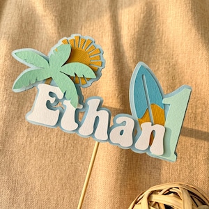 Peut inclure: Décoration de gâteau en papier superposé avec le nom "Ethan" en blanc, un palmier, un soleil, une planche de surf et le chiffre "1". La décoration est dans les tons de bleu, vert et jaune, fixée à un bâton en bois.