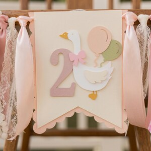 以下が含まれることがあります： 2歳の誕生日のお祝い用バナー。ピンクのリボンをつけた白いガチョウが2つの風船を持っています。数字の「2」はピンク色です。バナーはピンクのリボンとレースで飾られています。
