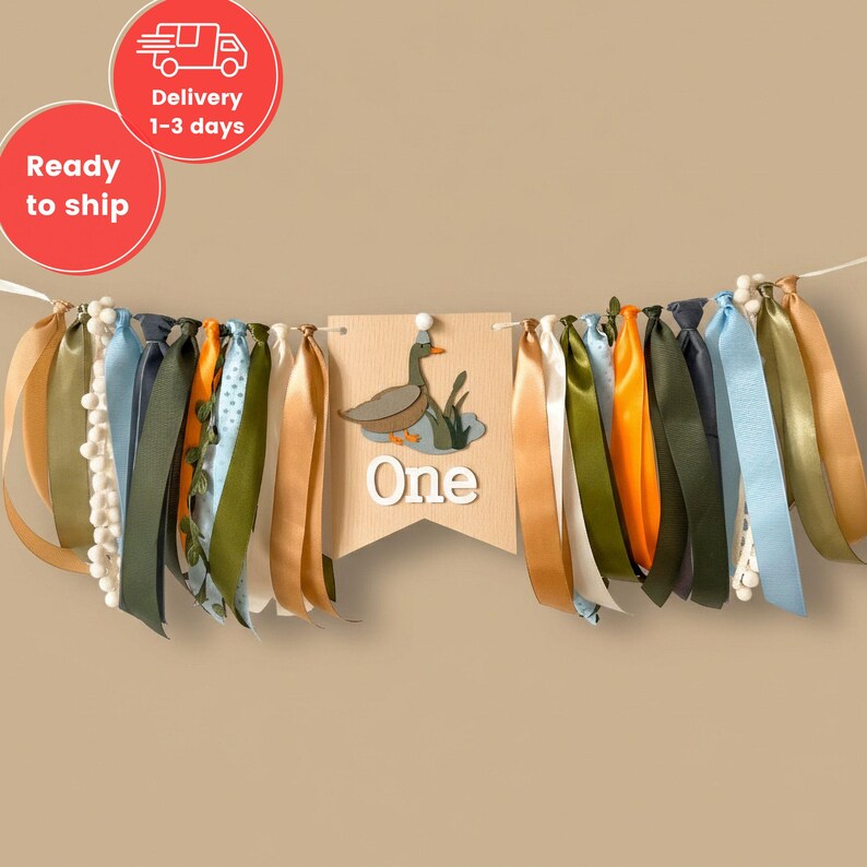 May include: A decorative banner with colorful ribbons and a wooden pennant. The pennant features a duck illustration and the word "One". Ribbons in shades of gold, blue, green, and orange are attached to the banner, creating a festive look.
