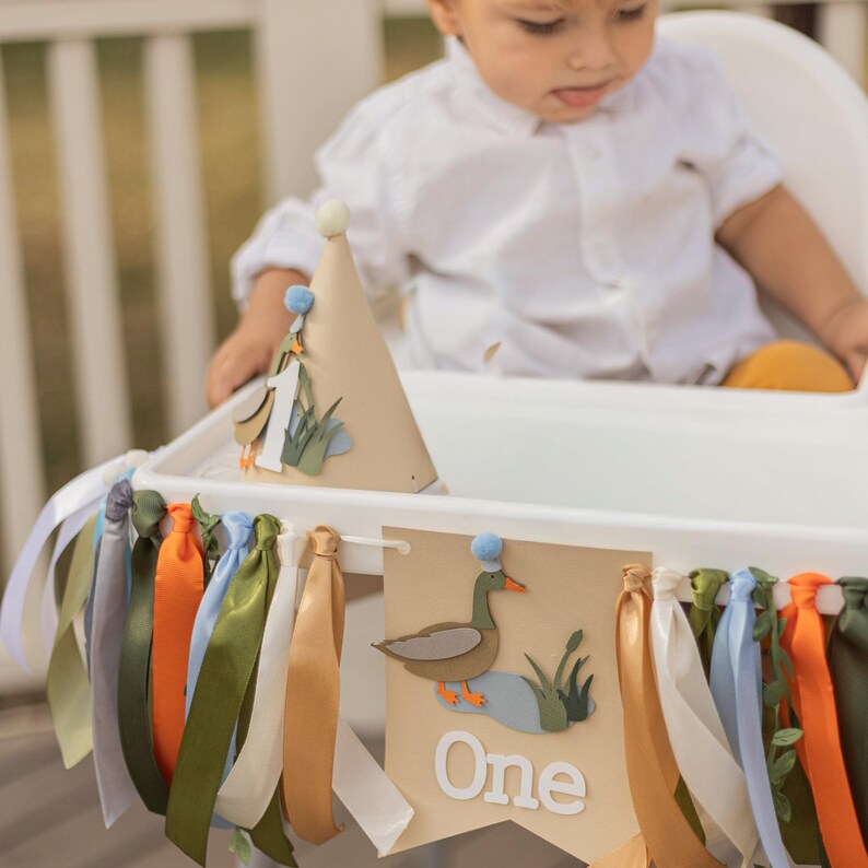 May include: A tan party hat with a duck and the number one, and a banner with the word "One" and a duck illustration. The banner is decorated with colorful ribbons. The theme is a first birthday celebration.