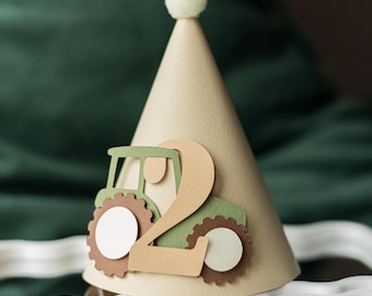 May include: A beige cone-shaped party hat featuring a layered paper cutout of a green tractor and the number two in a tan color. The hat is topped with a small, fuzzy, light-colored pom-pom and has a textured surface throughout the cone.