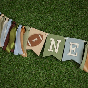 Puede incluir: Una pancarta verde, azul y marrón con la palabra "ONE" y una pelota de fútbol marrón en ella. La pancarta está hecha de fieltro y tiene una cuerda de cinta atada a ella.