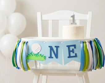 May include: A white high chair decorated with a light blue banner featuring a white golf ball, green grass, and the letters NE, accompanied by blue, green, and white ribbon tassels. A small white cake with a silver number one candle sits on top.