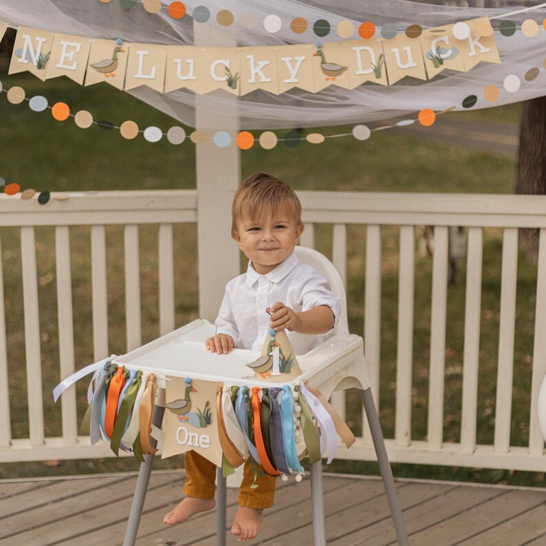 May include: A high chair decorated with a banner that says "One" and "Lucky Duck". The banner has duck illustrations and colorful ribbons. A child is sitting in the chair, wearing a white shirt and yellow pants.