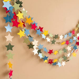 May include: A decorative garland featuring numerous five-pointed stars in shades of red, yellow, light blue, dark blue, olive green, and cream. The stars are connected by a thin white thread, creating a cascading, multi-layered hanging display.