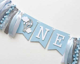 Könnte beinhalten: Ein hellblaues Banner mit dem Wort ONE in weißen Blockbuchstaben. Die erste Fahne zeigt eine graue Heißluftballon-Grafik mit weißen Wolken. Das Banner ist mit hängenden Bändern in Blau-, Weiß- und Grautönen verziert, darunter gepunktete Muster und hellblaue Pompon-Borten.