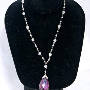 May include: A silver necklace with a teardrop-shaped, purple pendant. The necklace features a delicate chain with clear, round beads. The pendant is a deep purple color and is accented with silver detailing. The necklace is displayed on a black jewelry stand.