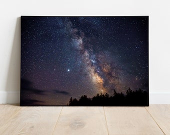 Puede incluir: Impresión en lienzo enmarcada que representa la Vía Láctea. La imagen muestra un cielo nocturno oscuro lleno de estrellas, con la Vía Láctea en el centro. Siluetas de árboles en la parte inferior crean un contraste llamativo.
