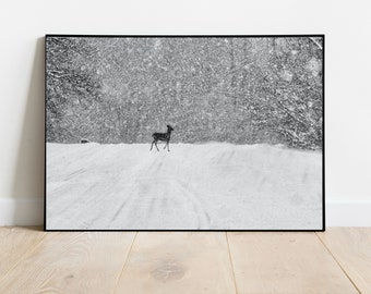 May include: Black and white framed print of a deer walking in a snowy landscape. The image captures a deer in silhouette against a backdrop of falling snow and a snow-covered hill. The artwork is framed in a black border.