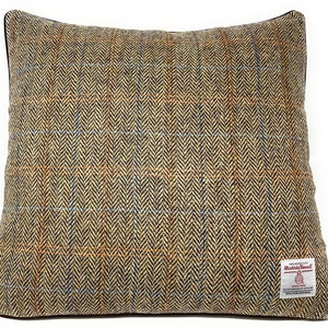Op de afbeelding: Een bruin, beige en blauw tweed kussen met een zwarte rand en een rood label met de tekst "Harris Tweed" met een klein logo van een schaap en een cirkel met de woorden "Handwoven" erin.