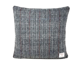 May include: A square throw pillow featuring a dark gray, blue, and white plaid pattern with thin vertical red stripes. The fabric has a textured, woven appearance. A small white rectangular label with black text is attached to the bottom right corner.