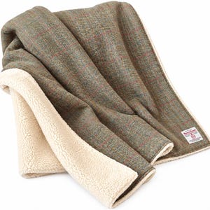 May include: A folded throw blanket featuring a green and brown herringbone tweed pattern with thin red and blue overcheck lines. The reverse side is lined with a cream-colored sherpa fleece fabric. A Harris Tweed label is sewn on the corner.
