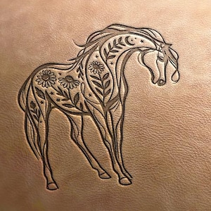 Pode incluir: Imagem gravada de um cavalo em uma superfície marrom. O cavalo apresenta desenhos florais e celestiais, incluindo flores, folhas, luas e estrelas. A crina e a cauda do cavalo fluem graciosamente, e o design é intrincado e artístico.