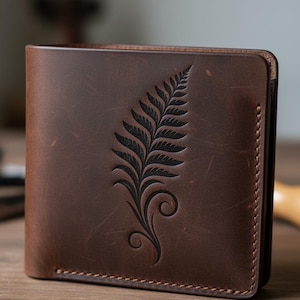 May include: Brown leather wallet with a fern leaf design. The wallet is made of a rich, textured leather and features a detailed, embossed fern on the front. The wallet is rectangular and appears to be bi-fold.