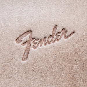Può includere: Primo piano di una superficie beige strutturata con la parola "Fender" in rilievo in una scrittura corsiva. Le lettere sono di una tonalità marrone più scura, creando un sottile contrasto con lo sfondo.
