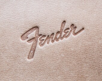 May include: Close-up of a textured, tan surface with the embossed word "Fender" in a cursive script. The lettering is a darker shade of brown, creating a subtle contrast against the background.