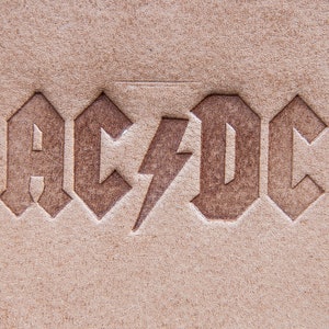 May include: A close-up of the word "AC/DC" embossed in brown on a light brown leather surface.