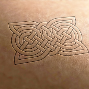 May include: A Celtic knot design carved into a brown leather surface. The knot is intricate and detailed, with a series of interwoven lines.