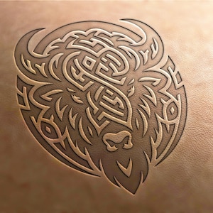 May include: A detailed, intricate line art design of a bison head, rendered in a tribal style. The design is carved into a light brown leather surface.