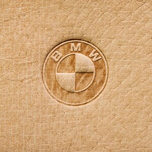 Può includere: Un primo piano di una superficie in pelle marrone con un logo BMW circolare in rilievo al centro. Il logo è di colore marrone chiaro e presenta le lettere "BMW" attorno all'esterno del cerchio.