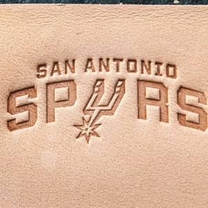 Może przedstawiać: Brązowa skórzana łatka z wytłoczonym napisem "San Antonio Spurs" brązową czcionką. Logo Spurs jest również wytłoczone w środku łatki.
