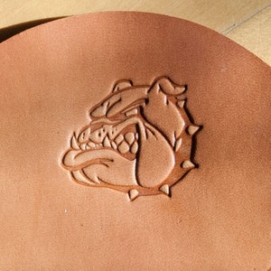 May include: A brown leather patch with a detailed embossed image of a bulldog's head. The bulldog has a fierce expression with sharp teeth and a furrowed brow.