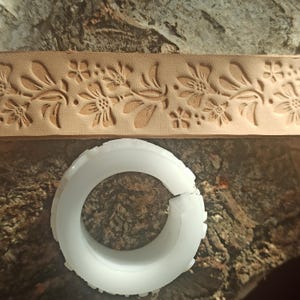 Puede incluir: Una tira de cuero marrón claro con un patrón floral y un objeto circular blanco con un borde texturizado. La tira de cuero presenta flores y hojas en relieve. El objeto blanco es un artículo en forma de anillo.
