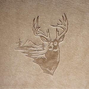 May include: A brown leather background with a carved image of a deer with large antlers. The deer is facing forward with its head tilted slightly to the right. The deer is surrounded by a carved image of mountains and trees.