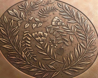 May include: A detailed, oval-shaped, bronze-colored decorative piece featuring an embossed floral design. The design includes various leaves, flowers, and berries, creating a natural, botanical theme. The piece has a textured surface.