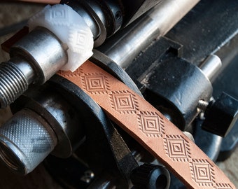 May include: A close-up of a leather stamping machine with a brown leather strap being embossed with a geometric pattern. The machine has a white plastic roller with a matching pattern.