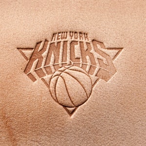 Può includere: Un cerotto in pelle marrone con un logo bianco in rilievo dei New York Knicks. Il logo presenta il nome della squadra "Knicks" in un font stilizzato con una palla da basket al centro.