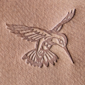 Puede incluir: Un fondo de cuero marrón con un diseño de colibrí blanco en relieve en la superficie. El colibrí está mirando hacia la derecha y tiene las alas extendidas.