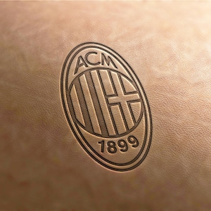 May include: A brown leather surface with an embossed oval logo. The logo features the letters "ACM" above a striped design and a cross, with the number "1899" below. The overall design is a classic emblem.