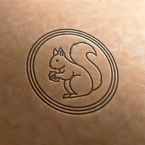 May include: A circular emblem featuring a detailed outline of a squirrel holding a nut, set within a triple-lined border. The design is embossed on a light brown leather surface, creating a textured appearance.