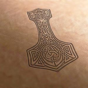 Puede incluir: Imagen detallada en marrón oscuro del martillo de Thor, Mjolnir, tallado en una superficie de cuero marrón claro. El martillo presenta intrincados diseños de nudos y geométricos, con un prominente emblema circular en la base.