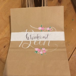 May include: Brown paper gift bags with twisted paper handles. Each bag features a white band with the text "b.tricker.art" in black script and white cursive. Pink and green floral accents are above and below the text.