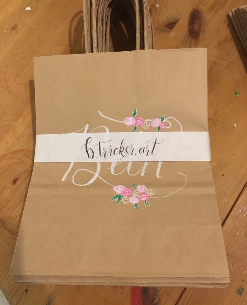 May include: Brown paper gift bags with twisted paper handles. Each bag features a white band with the text "b.tricker.art" in black script and white cursive. Pink and green floral accents are above and below the text.