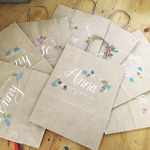 May include: A collection of kraft paper gift bags with white calligraphy names and floral designs in various colors. Each bag has a matching paper handle and the date "31st May 2019" is written on some of the bags.