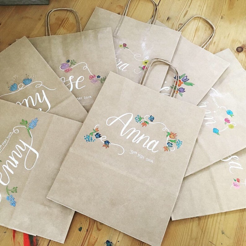 May include: A collection of kraft paper gift bags with white calligraphy names and floral designs in various colors. Each bag has a matching paper handle and the date "31st May 2019" is written on some of the bags.