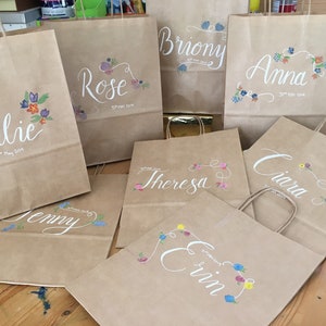 May include: Kraft paper gift bags with names in elegant white script and floral accents. Each bag features a different name, such as "Julie," "Rose," and "Anna," along with the date "31st May 2019."
