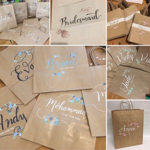 May include: A collection of brown paper gift bags with handwritten names and floral accents. Some bags say "Bridesmaid" or "Just for You". The names are in various colors, with blue and pink floral designs.