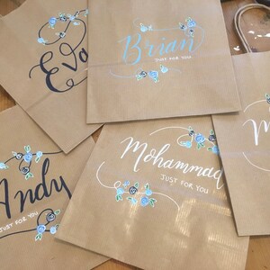 May include: Kraft paper gift bags with names written in calligraphy. Each bag features a name, such as "Eva", "Brian", "Andy", "Mohammad", and "Michael", with "Just For You" below. Floral accents in blue and black decorate each bag.