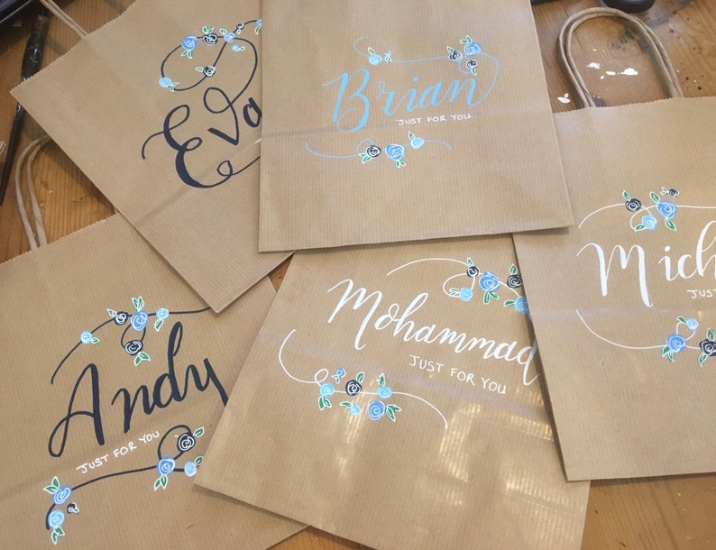 May include: Kraft paper gift bags with names written in calligraphy. Each bag features a name, such as "Eva", "Brian", "Andy", "Mohammad", and "Michael", with "Just For You" below. Floral accents in blue and black decorate each bag.