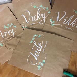 May include: Several brown paper gift bags with white calligraphy names and titles, including "Amy Maid of Honour", "Vicky Bridesmaid", "Vikki Bridesmaid", and "Jade chief bridesmaid". Each bag features a floral design in light green.