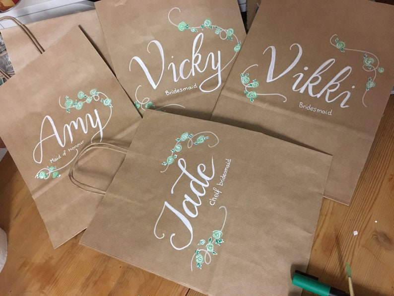 May include: Several brown paper gift bags with white calligraphy names and titles, including "Amy Maid of Honour", "Vicky Bridesmaid", "Vikki Bridesmaid", and "Jade chief bridesmaid". Each bag features a floral design in light green.