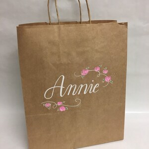 May include: A brown paper shopping bag with twisted paper handles. The word "Annie" is written in white script, with pink floral accents. The bag is designed for gift giving or retail use.